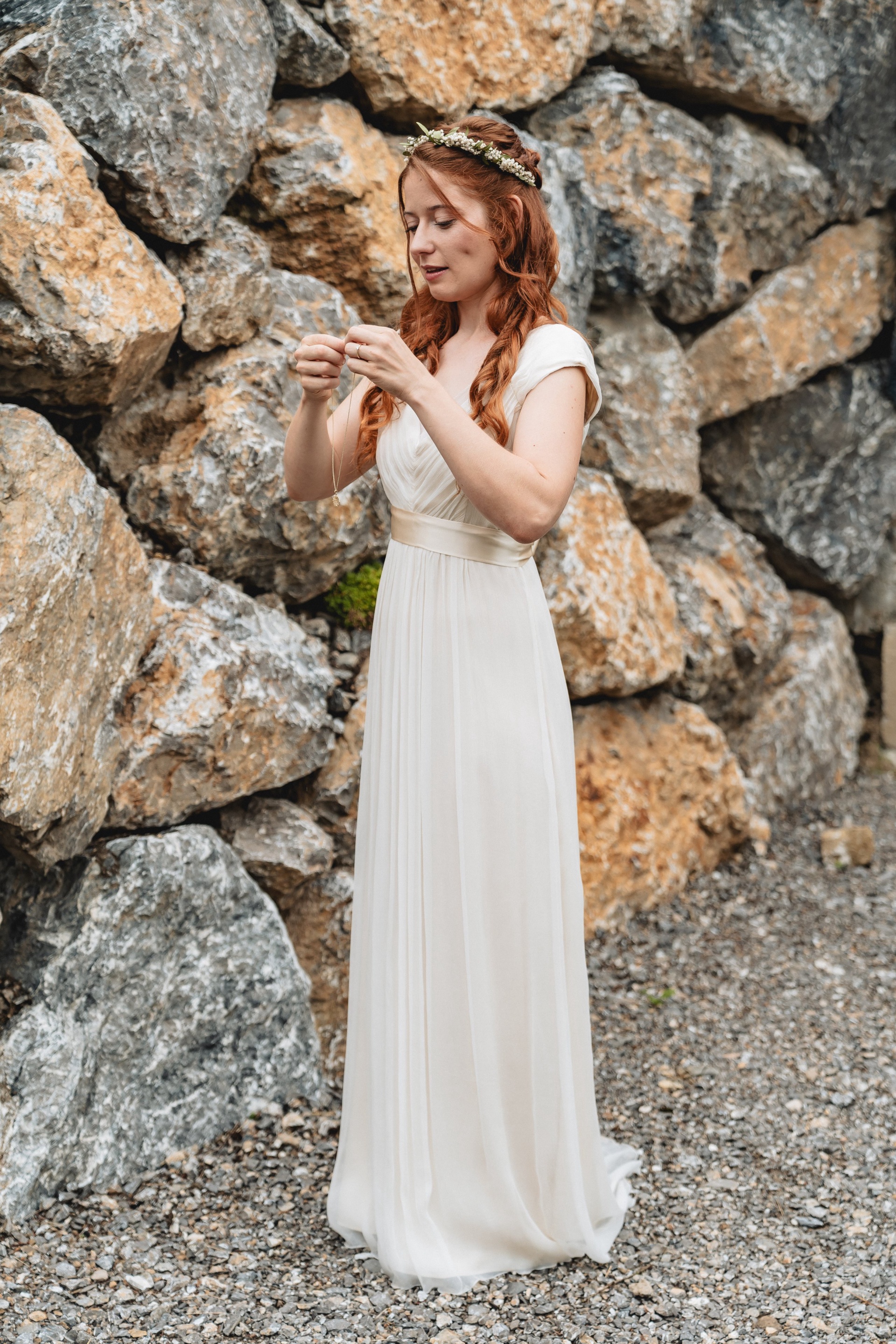 Braut Hannah im schlichten weißen Kleid mit Blumenkranz vor der Steinmauer